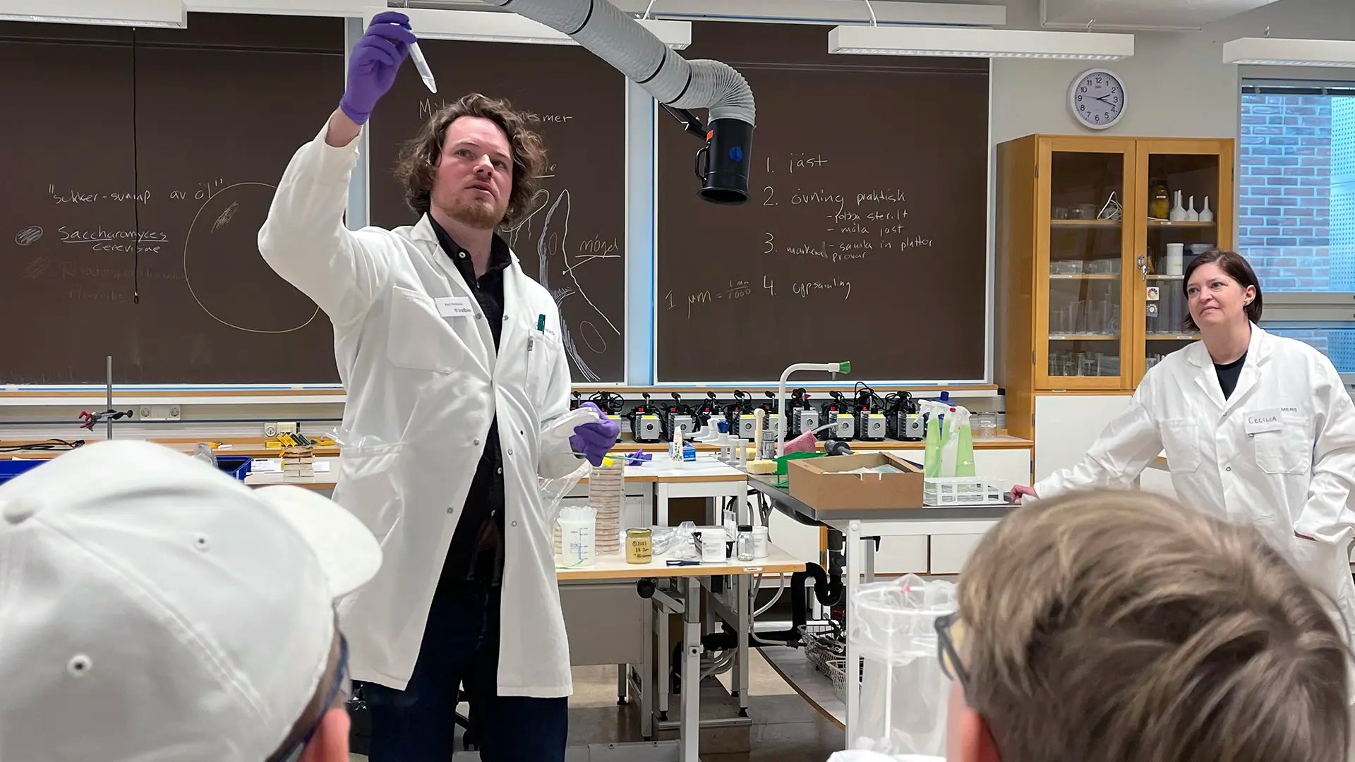 Två forskare i labbrockar visar labbutrustning