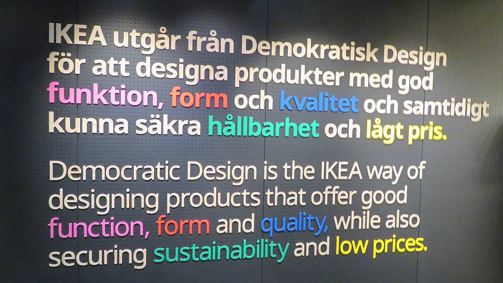Vit text på en svart vägg som beskriver IKEAs grundpelare för design. Dessa ord skrivs i olika färger.