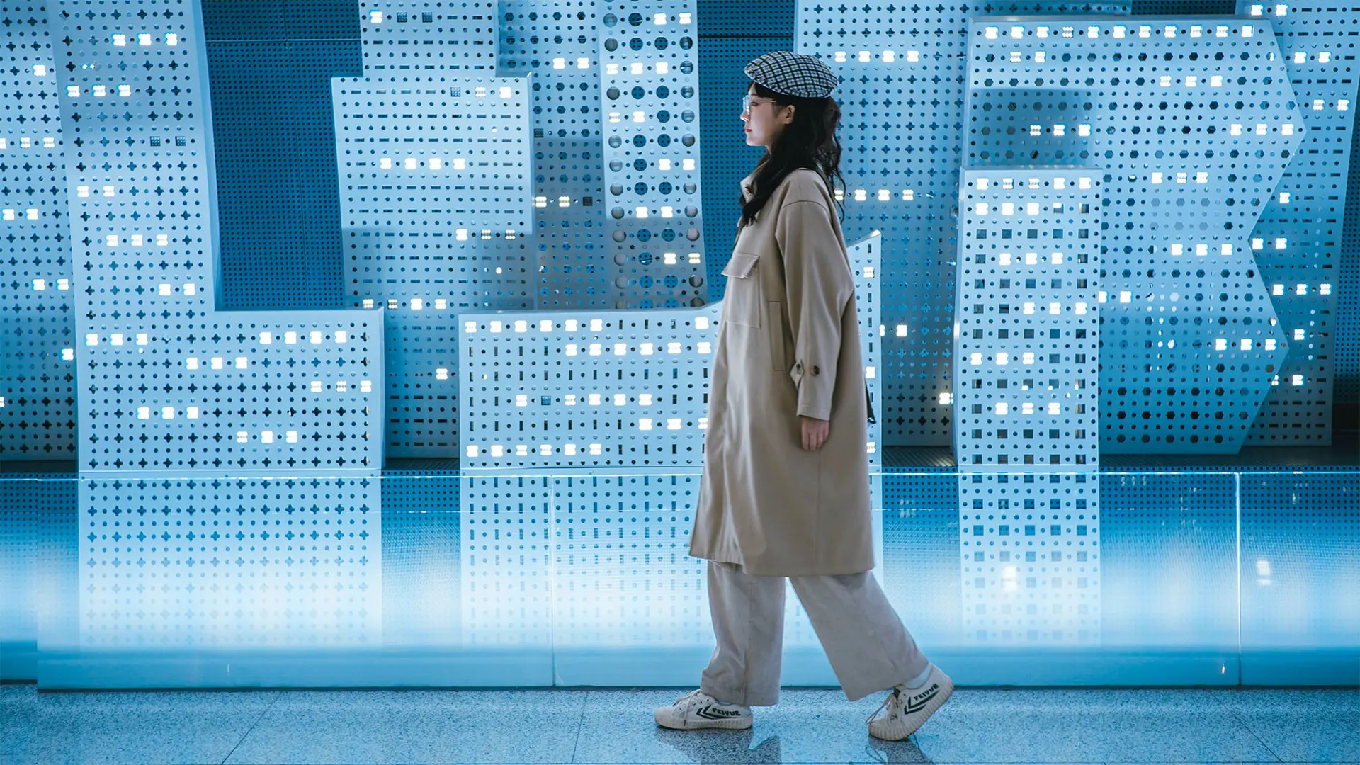 Woman walking in front of background of tiles with holes and lights reminiscent of skyscrapers
